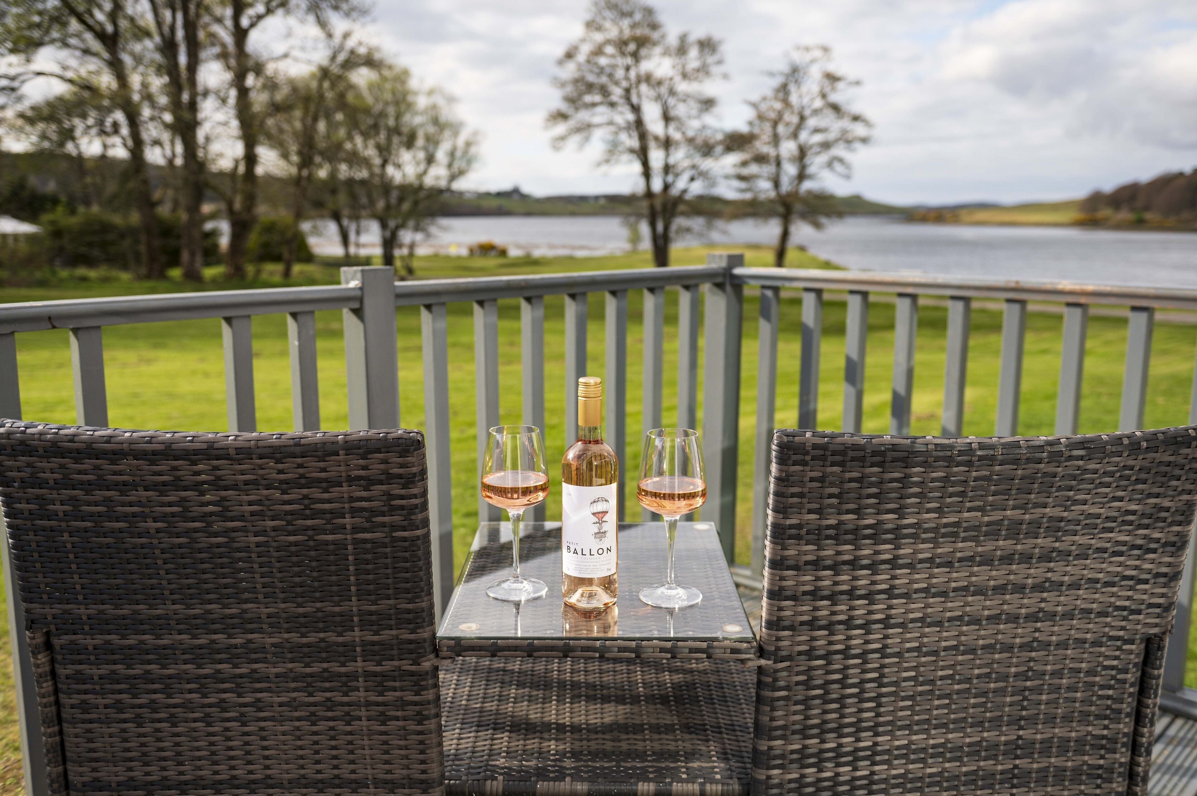 Une chaise et une table en osier, avec une bouteille et deux verres de vin, sur un balcon donnant sur un lac.