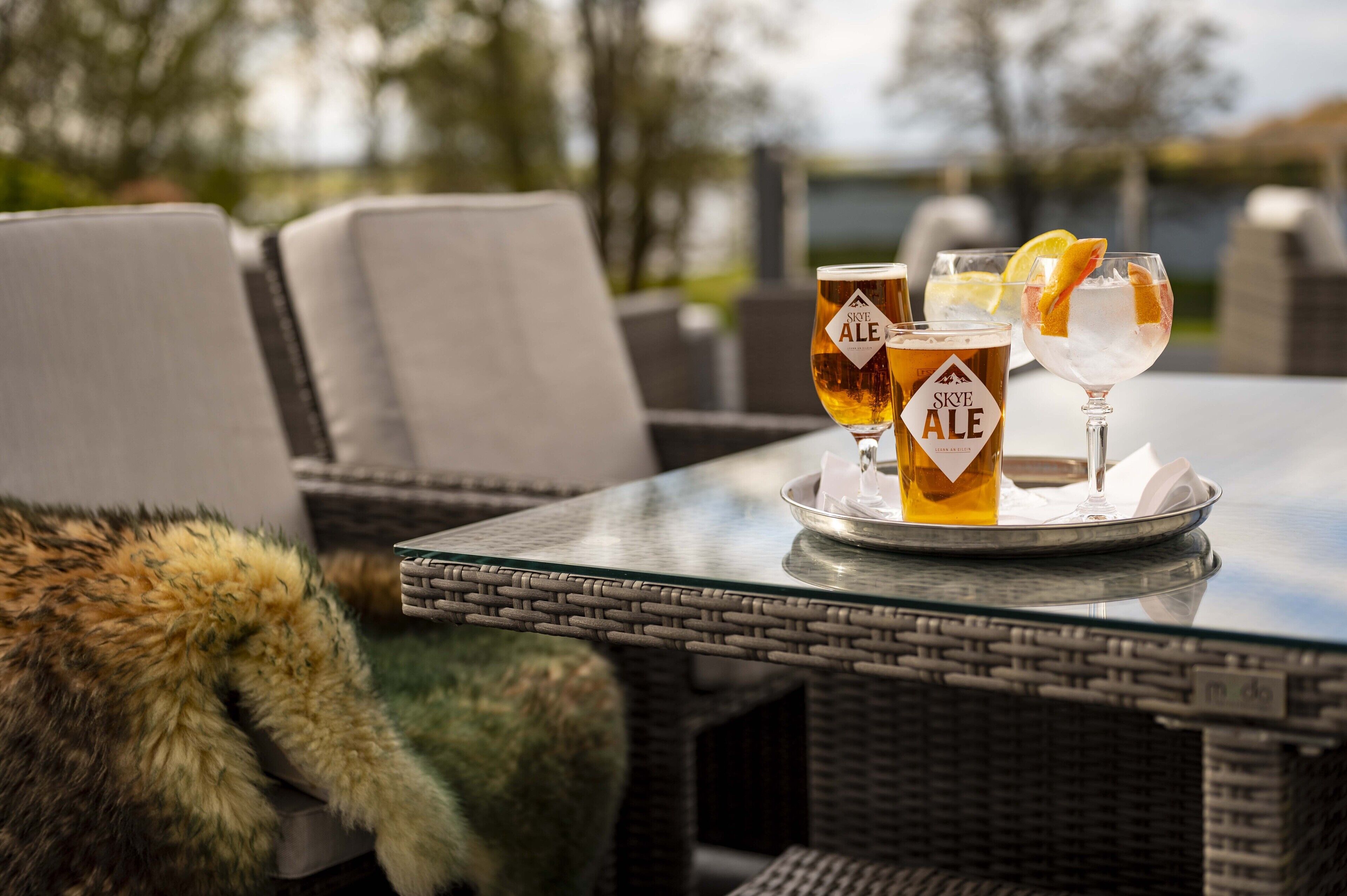 Una mesa con dos vasos de Skye Ale y un vaso de bebida helada, con una silla de mimbre y un plaid de piel en primer plano.