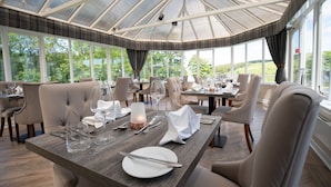 Un restaurant doté d’un haut plafond en verre et de grandes fenêtres offrant une vue sur l’extérieur. Les tables sont dressées avec des nappes blanches, des verres à vin et des couverts.