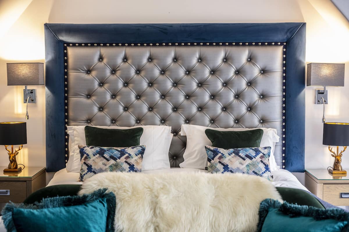 A bedroom with a tufted headboard, white and patterned pillows, a fluffy white throw, and two bedside tables with lamps and a decorative item.