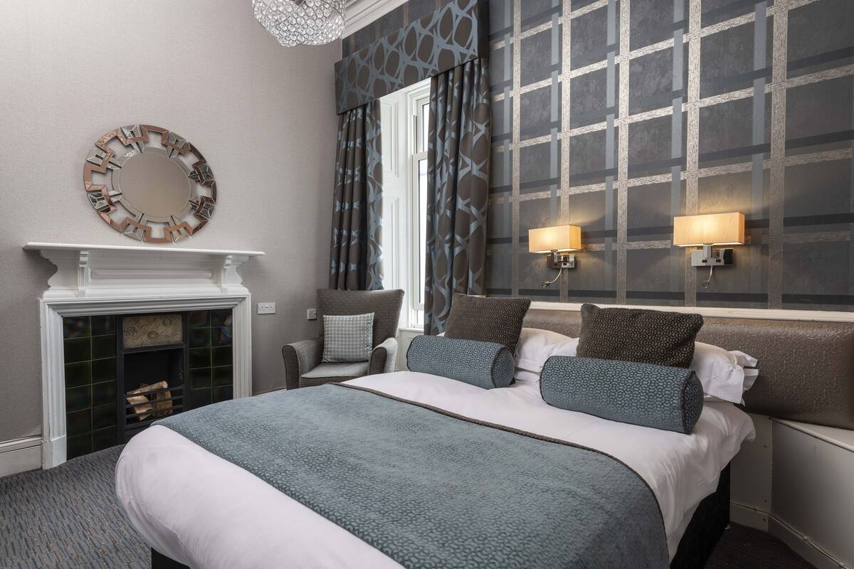 A hotel room with a large bed, a fireplace, two wall-mounted lamps, and a decorative mirror.