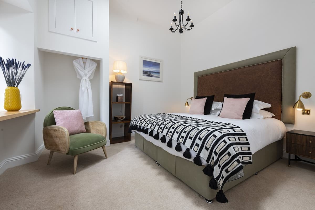 A bedroom with a large bed, a green armchair, a bedside table with a lamp, and a wall-mounted cabinet.