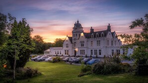 A large, historic building with a symmetrical facade, multiple windows, and a prominent tower. There is a well-maintained lawn in the foreground with several parked cars. - Skeabost House Hotel (Skeabost)