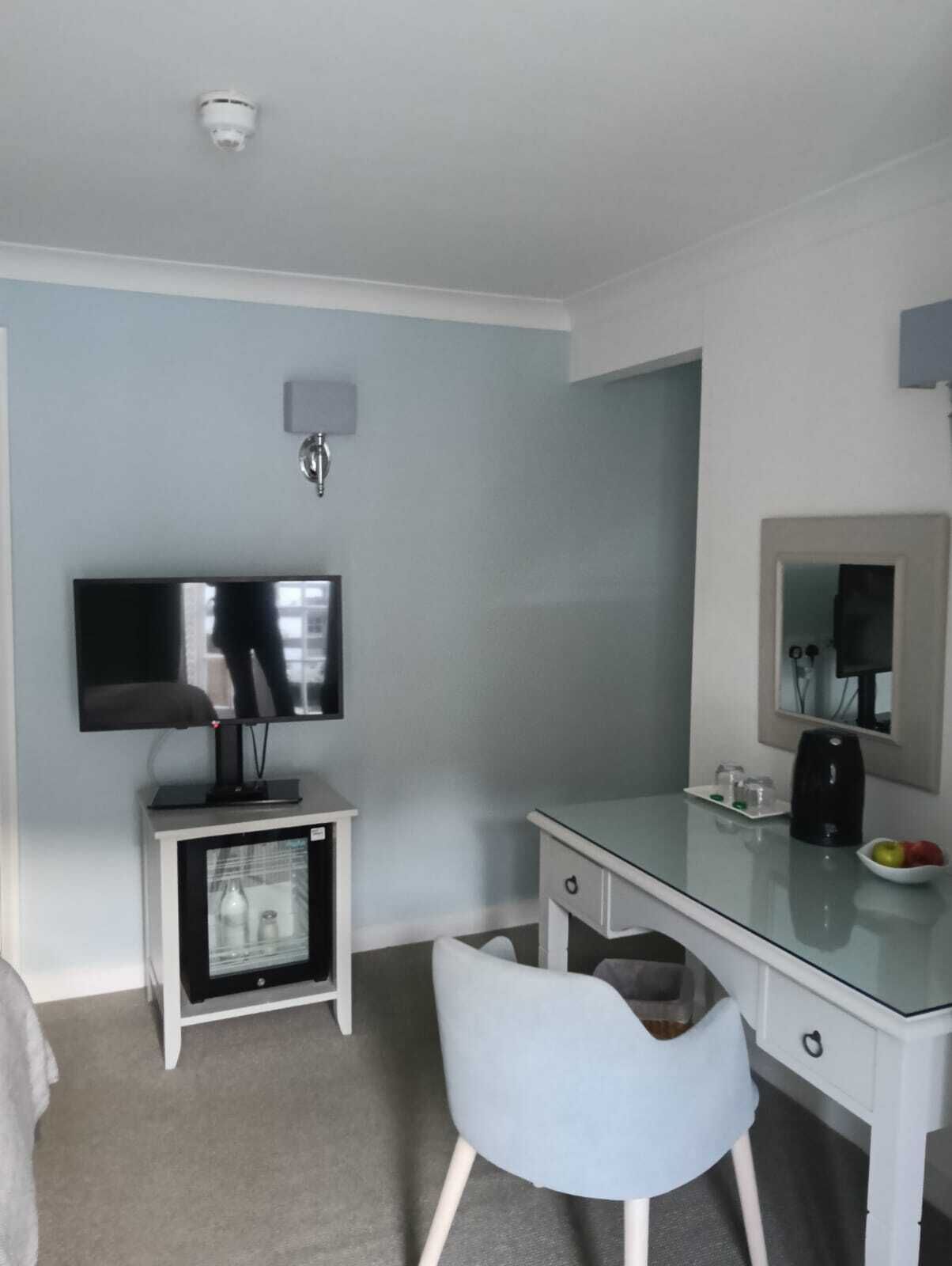 Habitación de hotel con televisión de pantalla plana sobre un soporte, un refrigerador pequeño, un escritorio de vidrio con cafetera, una silla y un espejo en la pared.