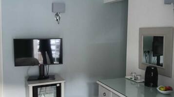 Habitación de hotel con televisión de pantalla plana sobre un soporte, un refrigerador pequeño, un escritorio de vidrio con cafetera, una silla y un espejo en la pared.