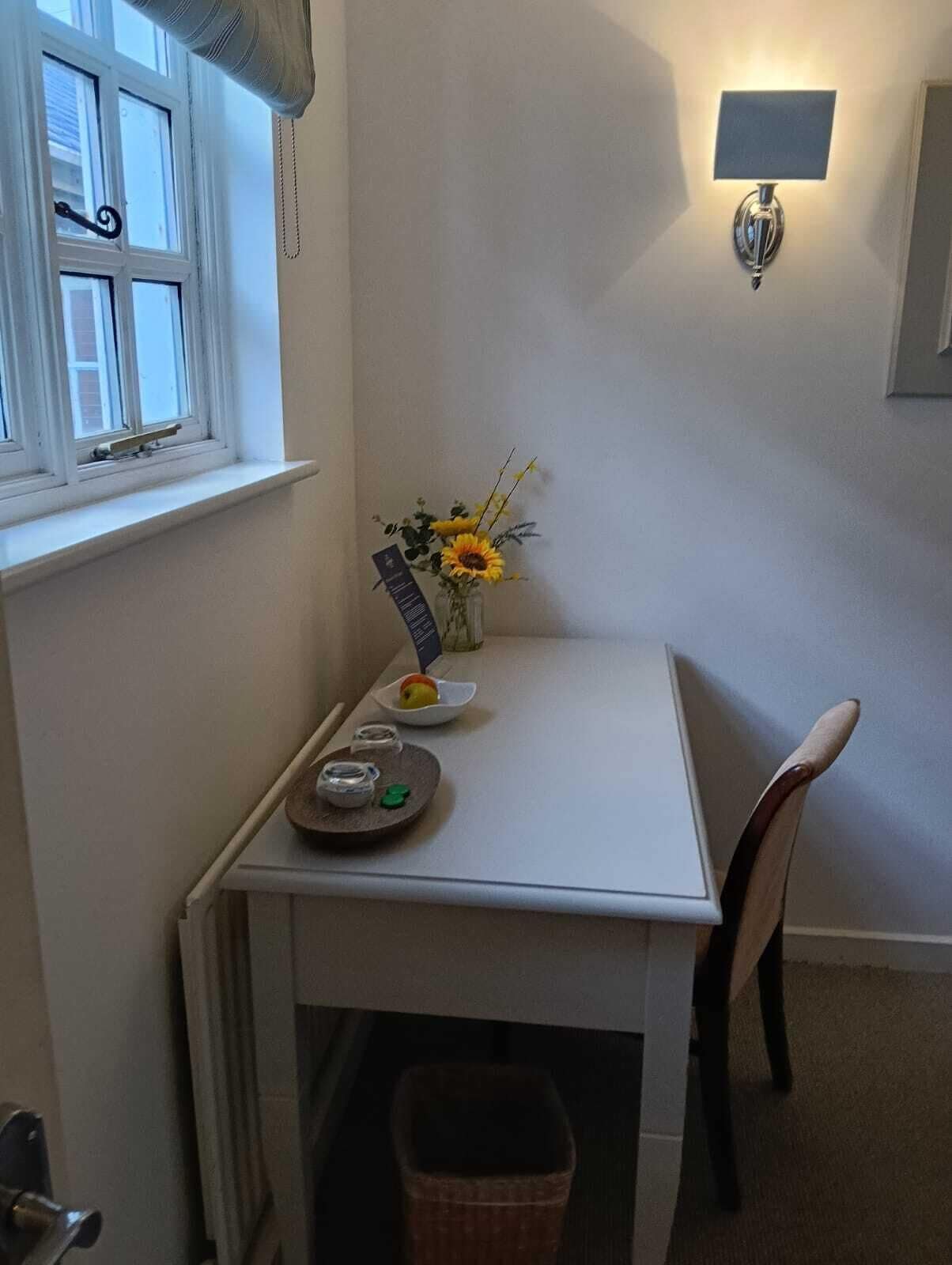 A small, white dining table with a chair, a vase with sunflowers, a small plate with an apple, and a tray with a teapot and cups.