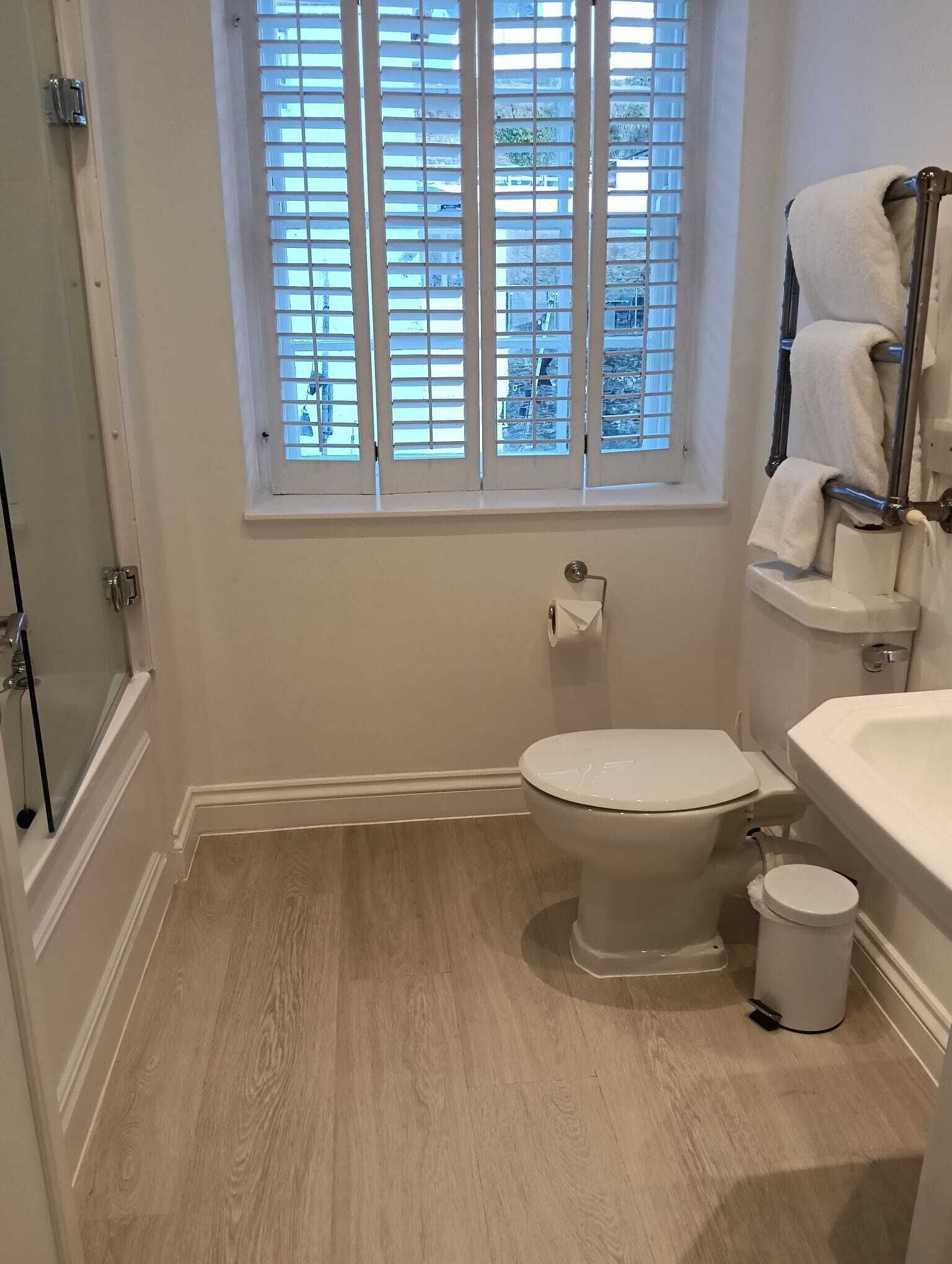 A bathroom with a toilet, a sink, a window with blinds, and a shower area.