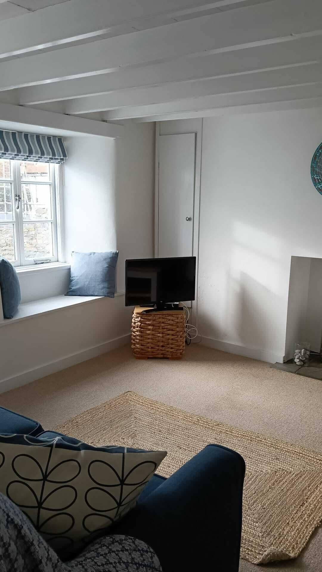 A living room with a television on a woven stool, a window with blue cushions, and a large woven rug on the floor.