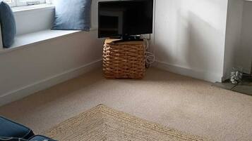 A living room with a television on a woven stool, a window with blue cushions, and a large woven rug on the floor.