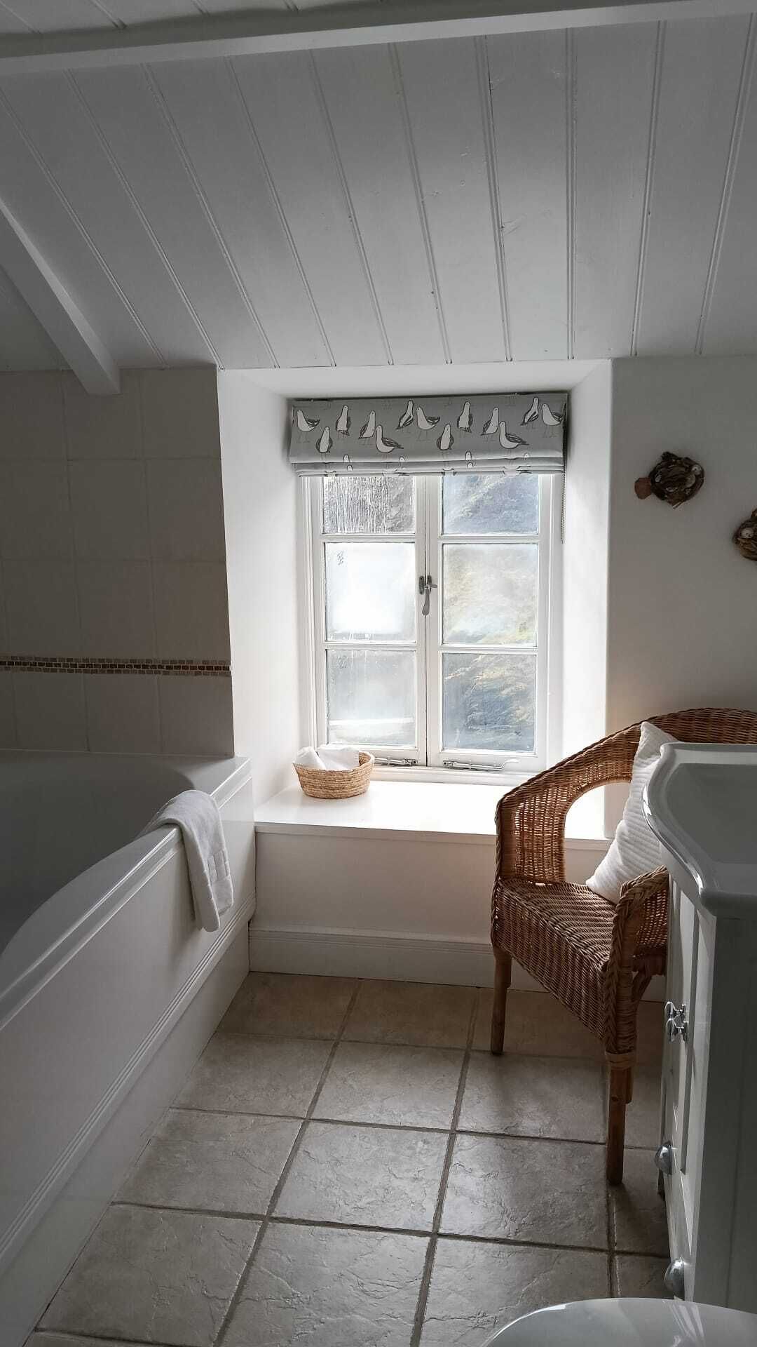 A bathroom with a bathtub, a wicker chair, a window with frosted glass, and a tiled floor.