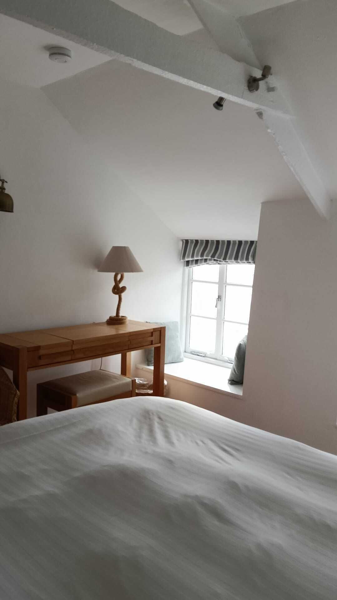 A bedroom with a bed, a wooden desk, a lamp, a window with a striped valance, and a ceiling light fixture.
