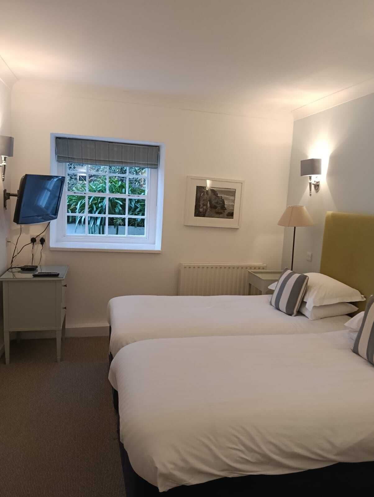 A hotel room with two beds, a television on a desk, a window with blinds, a wall-mounted lamp, and a framed picture.