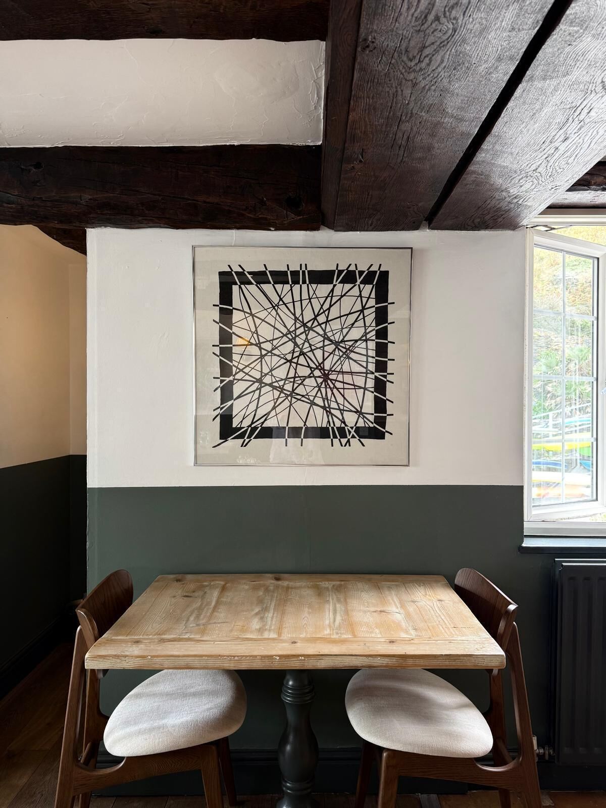 Un comedor con una mesa y sillas de madera, una obra de arte abstracta en la pared y una ventana con vistas a la vegetación.