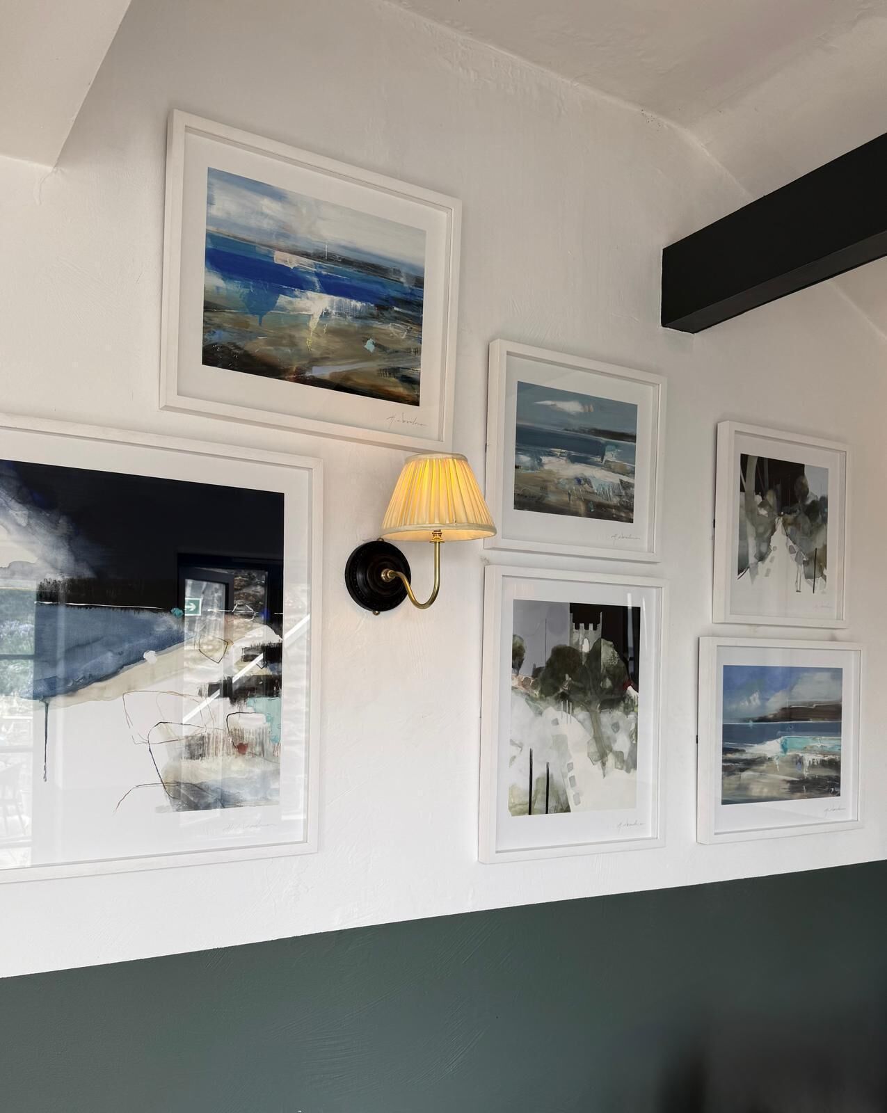A wall with multiple framed artworks, a wall-mounted lamp, and a dark-colored beam.