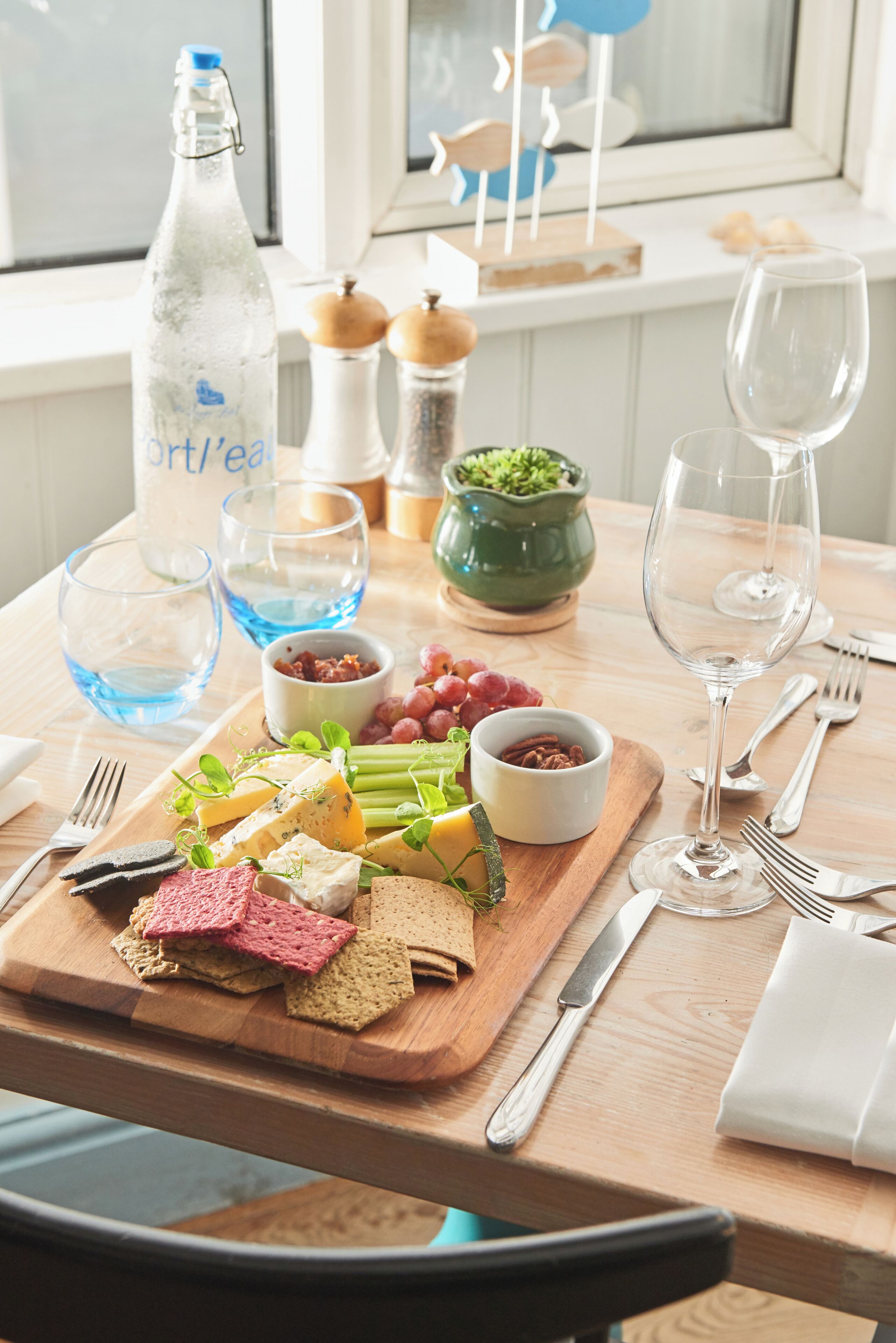 Una mesa de madera con una variedad de quesos, galletas y uvas, junto con una botella de agua y copas de vino.