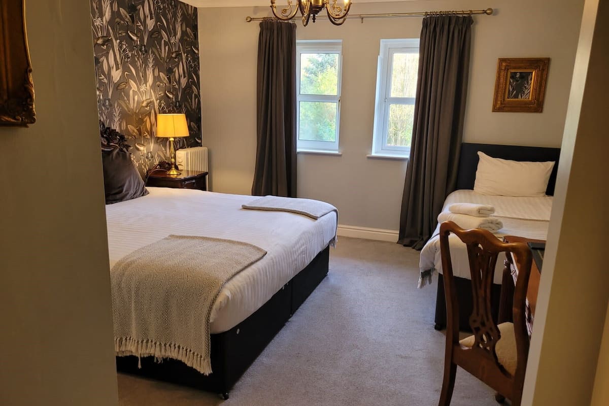 A bedroom with a large bed, a smaller bed, a chair, a nightstand with a lamp, and a wall with a floral pattern.