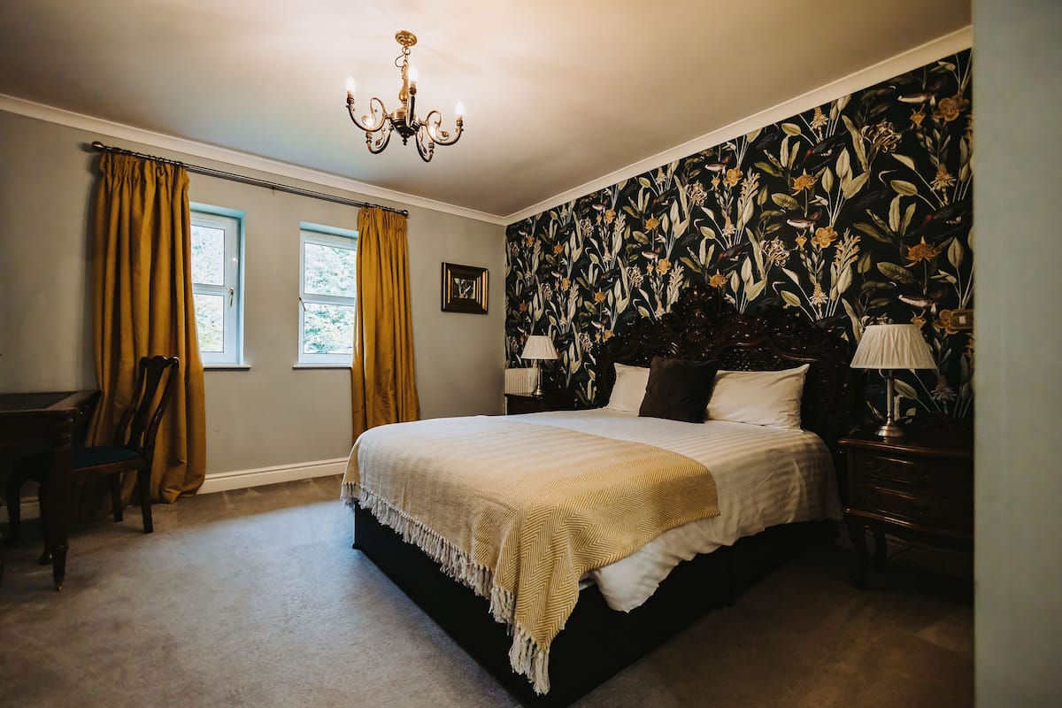 A bedroom with a large bed, two bedside tables with lamps, a chandelier, a window with curtains, and a floral wallpaper.