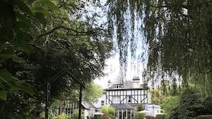 Um edifício tradicional em estilo Tudor, com fachada simétrica, um jardim bem cuidado com sebes e um gramado central, e um caminho de pedra que leva à entrada.