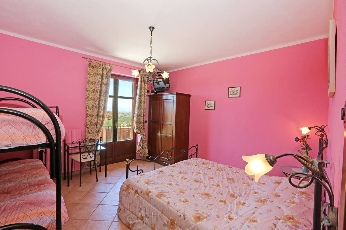 Un dormitorio con paredes rosas, una cama con cubrecama floral, un escritorio con silla, un guardarropa y una araña.
