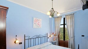 A bedroom with a bed, a small table, a wall-mounted lamp, a window with curtains, and a view of greenery outside. - Al Brich (Albaretto della Torre)
