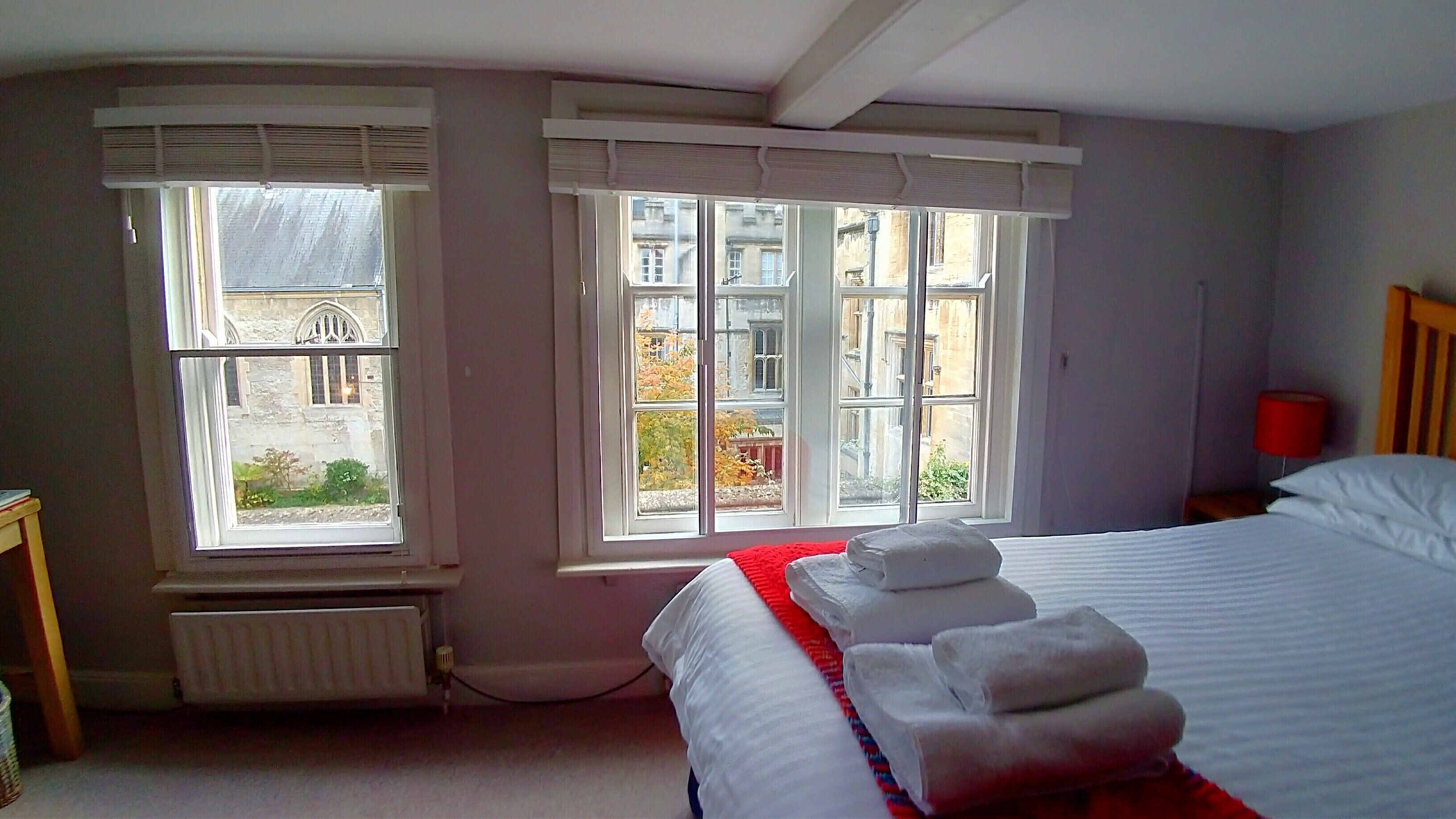 A hotel room with two large windows, a bed with white linens, and a red throw blanket.