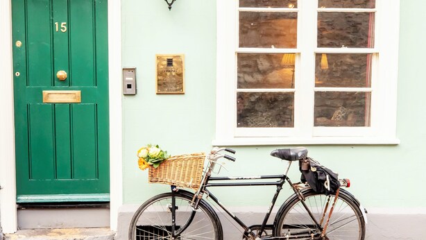 Una puerta verde con el número 15, una lámpara de pared, una ventana con cortinas y una bicicleta con cesta.