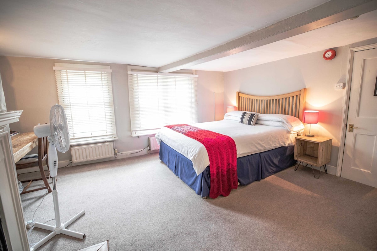 A bedroom with a bed, bedside table, lamp, and a standing fan.