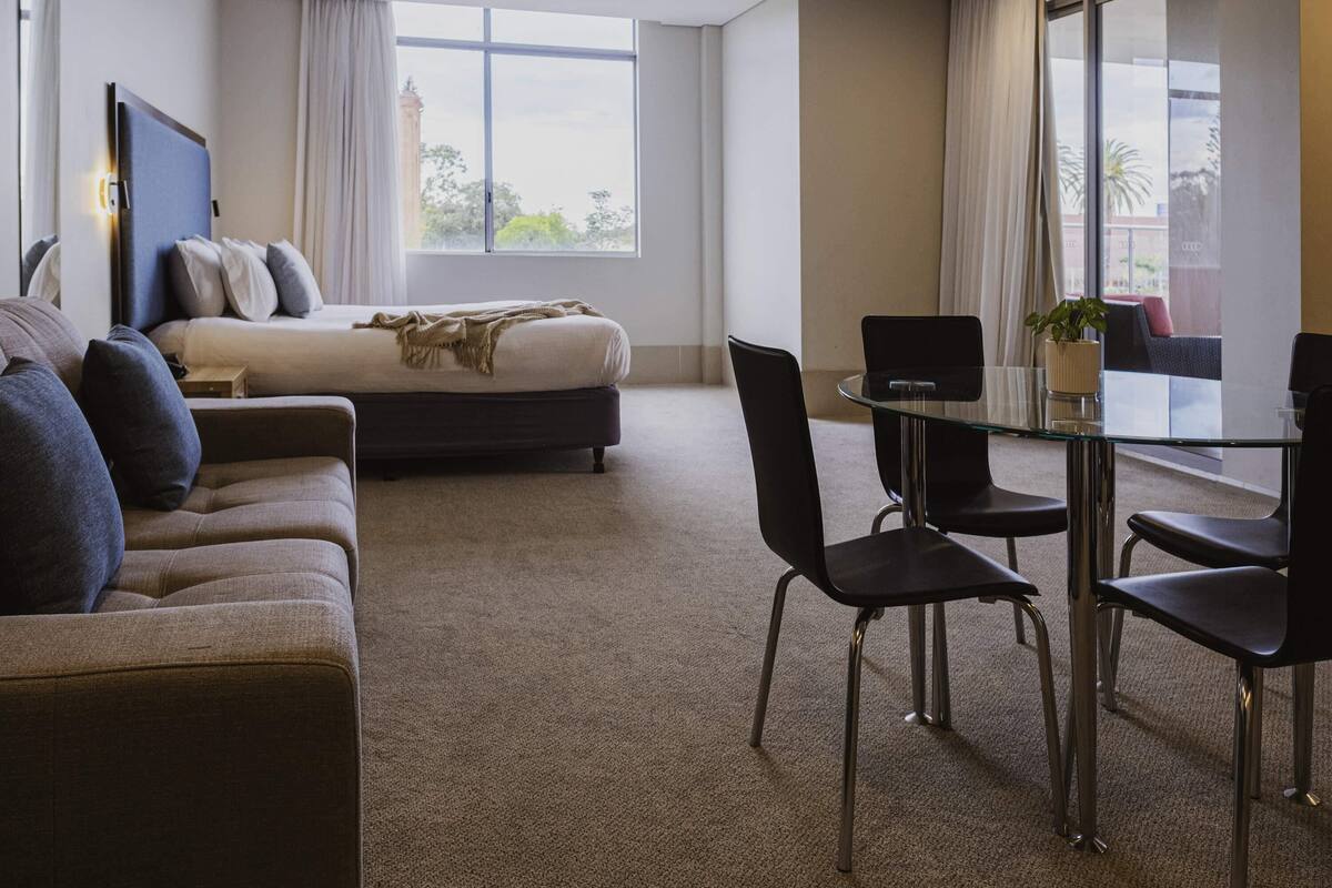 A modern hotel room with a bed, sofa, dining table, chairs, and large windows.