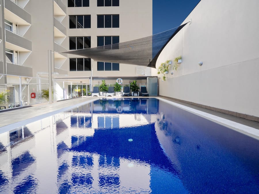 A swimming pool with clear water, surrounded by a modern building with balconies and a covered seating area.