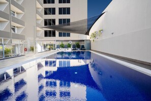 En swimmingpool med klart vatten, omgiven av en modern byggnad med balkonger och ett takförsett sittområde.