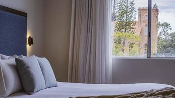 A bedroom with a bed, a window with curtains, a view of a building, and a wall-mounted light fixture.