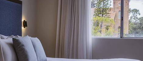 Un dormitorio con una cama, una ventana con cortinas, vistas a un edificio y una lámpara fijada a la pared.