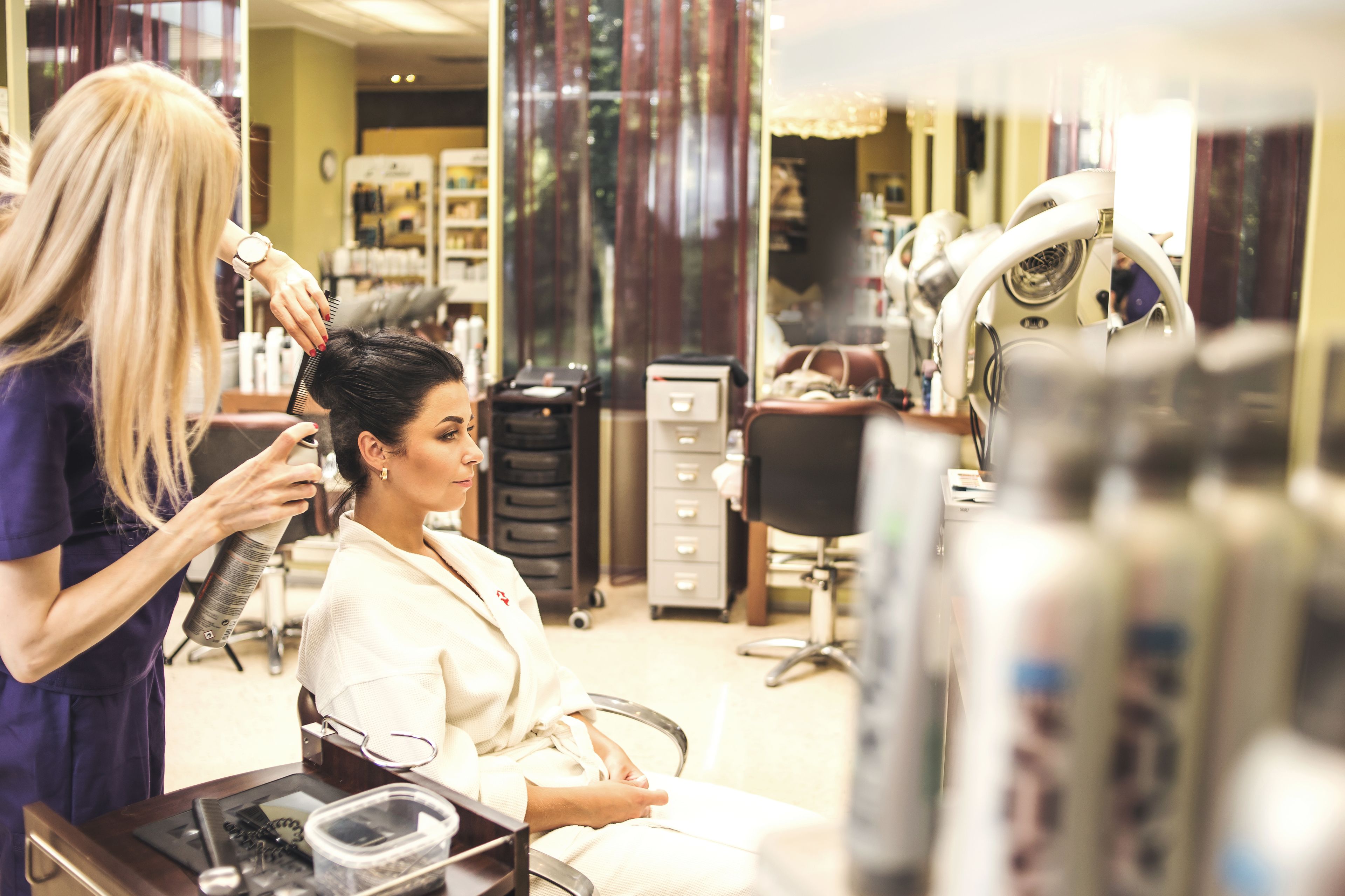 A salon interior with a client seated in a salon chair, a stylist attending to the client, and various salon equipment in the background.