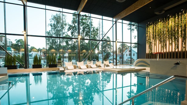 An indoor swimming pool with clear glass walls, lounge chairs, and a view of trees outside.