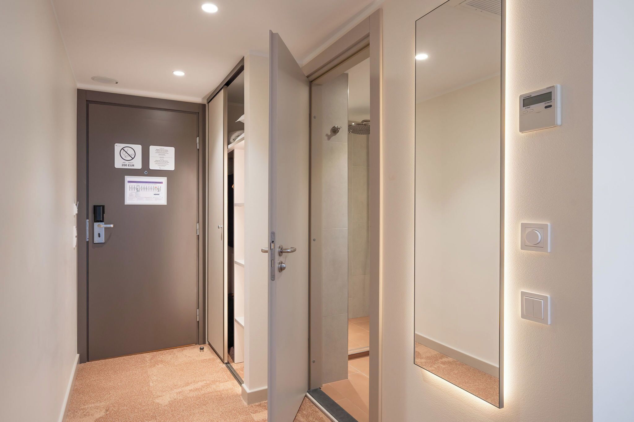 A hallway with a door leading to a bathroom, a shower, and a control panel on the wall.