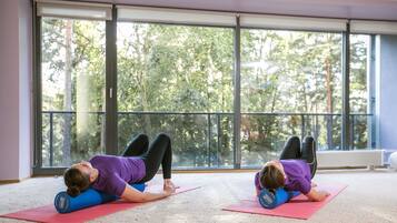 Two individuals performing a back stretch on yoga mats in a room with large windows and a view of trees.