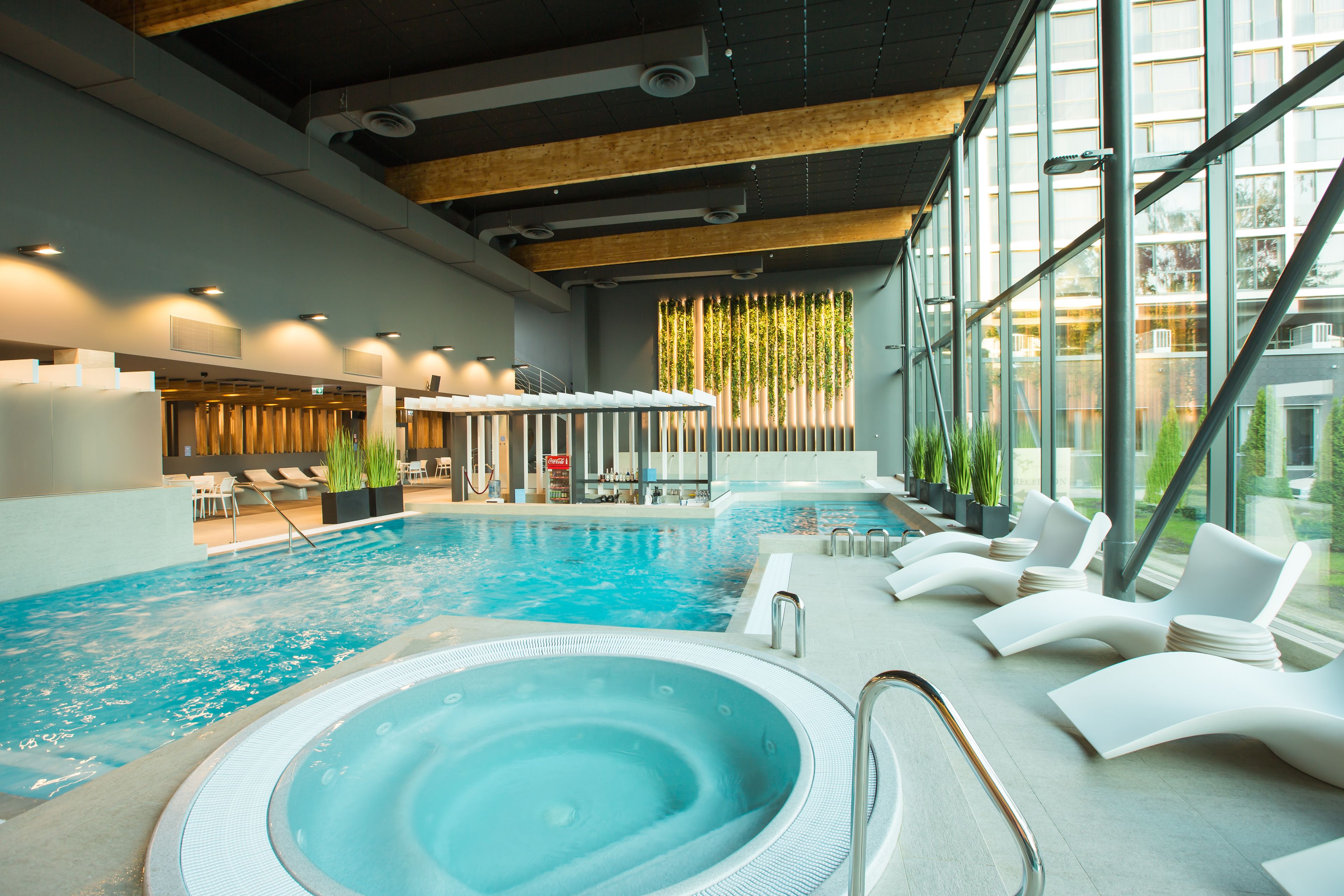 An indoor swimming pool with a hot tub, lounge chairs, and a view of the outside through large windows.