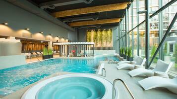 An indoor swimming pool with a hot tub, lounge chairs, and a view of the outside through large windows.