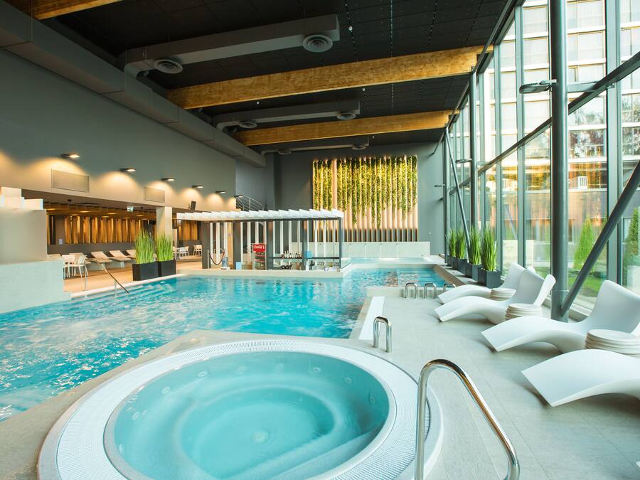 An indoor swimming pool with a hot tub, lounge chairs, and a view of the outside through large windows.