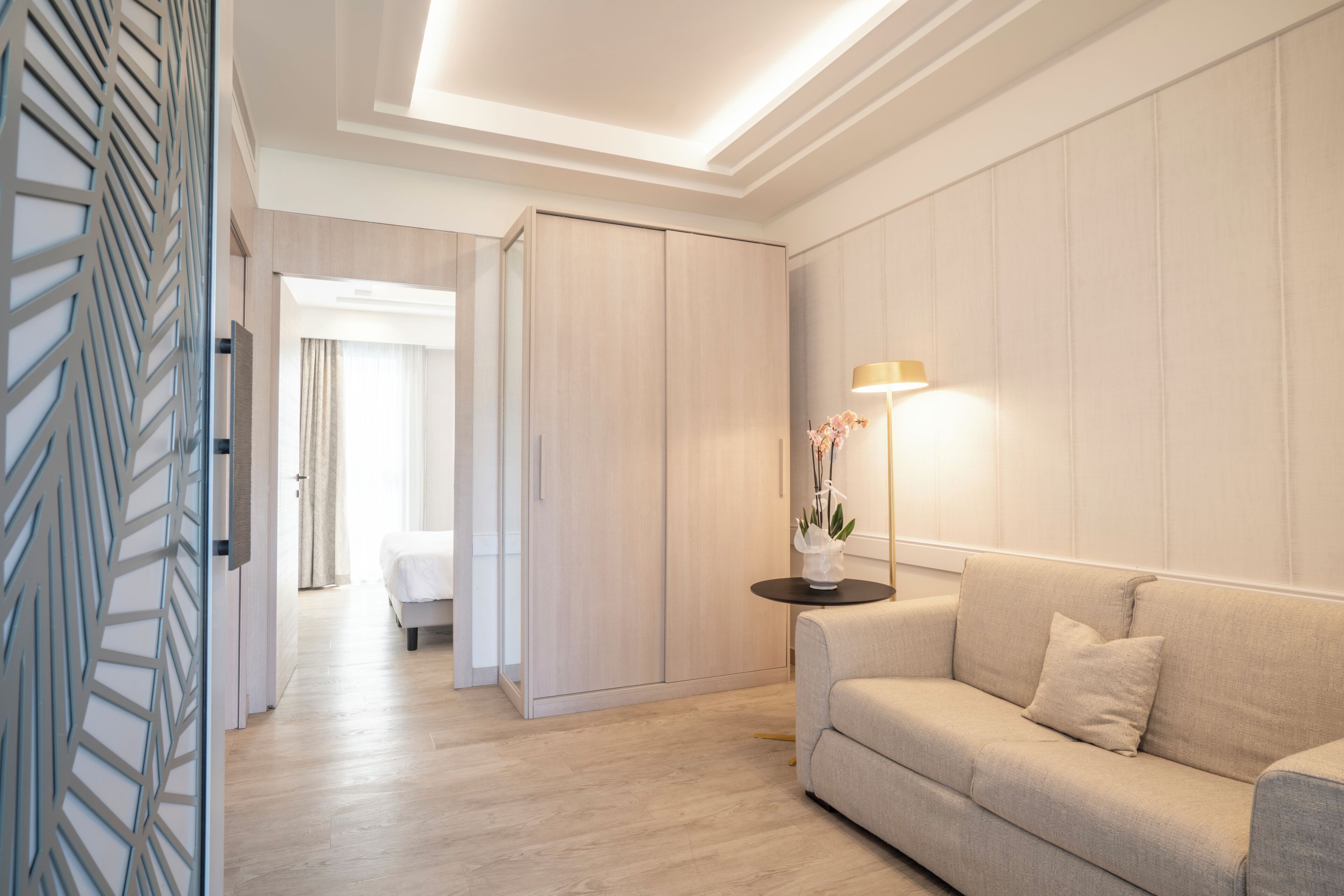 Et moderne hotelværelse med en sofa, et natbord med lampe, et garderobeskab og en dekorativ vægpanel.