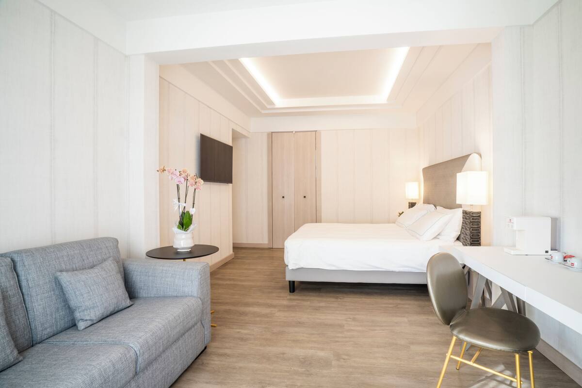 A modern hotel room with a grey sofa, a bed, a desk, and a chair.