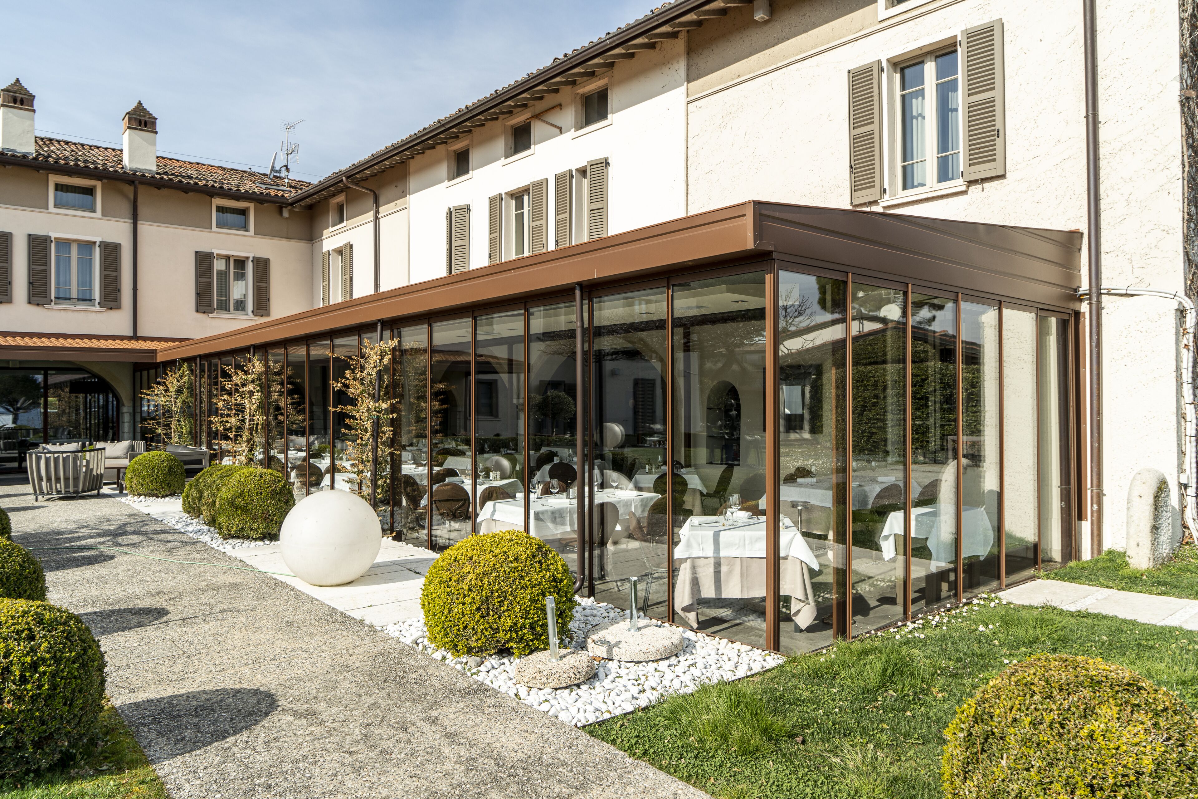 Un ristorante con posti a sedere all'aperto, pareti in vetro e un giardino curato con siepi ordinate e una grande scultura sferica.