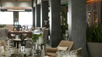 Una mesa con dispensador de agua, vasos y un plato en un interior moderno con sillas y plantas.