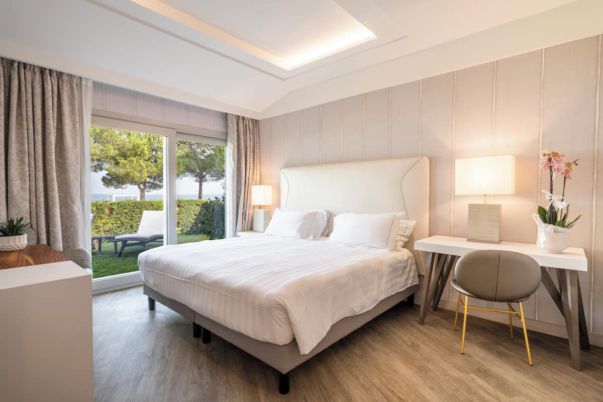 A hotel room with a large bed, a desk, and a chair. There is a view of the outdoors through the windows.