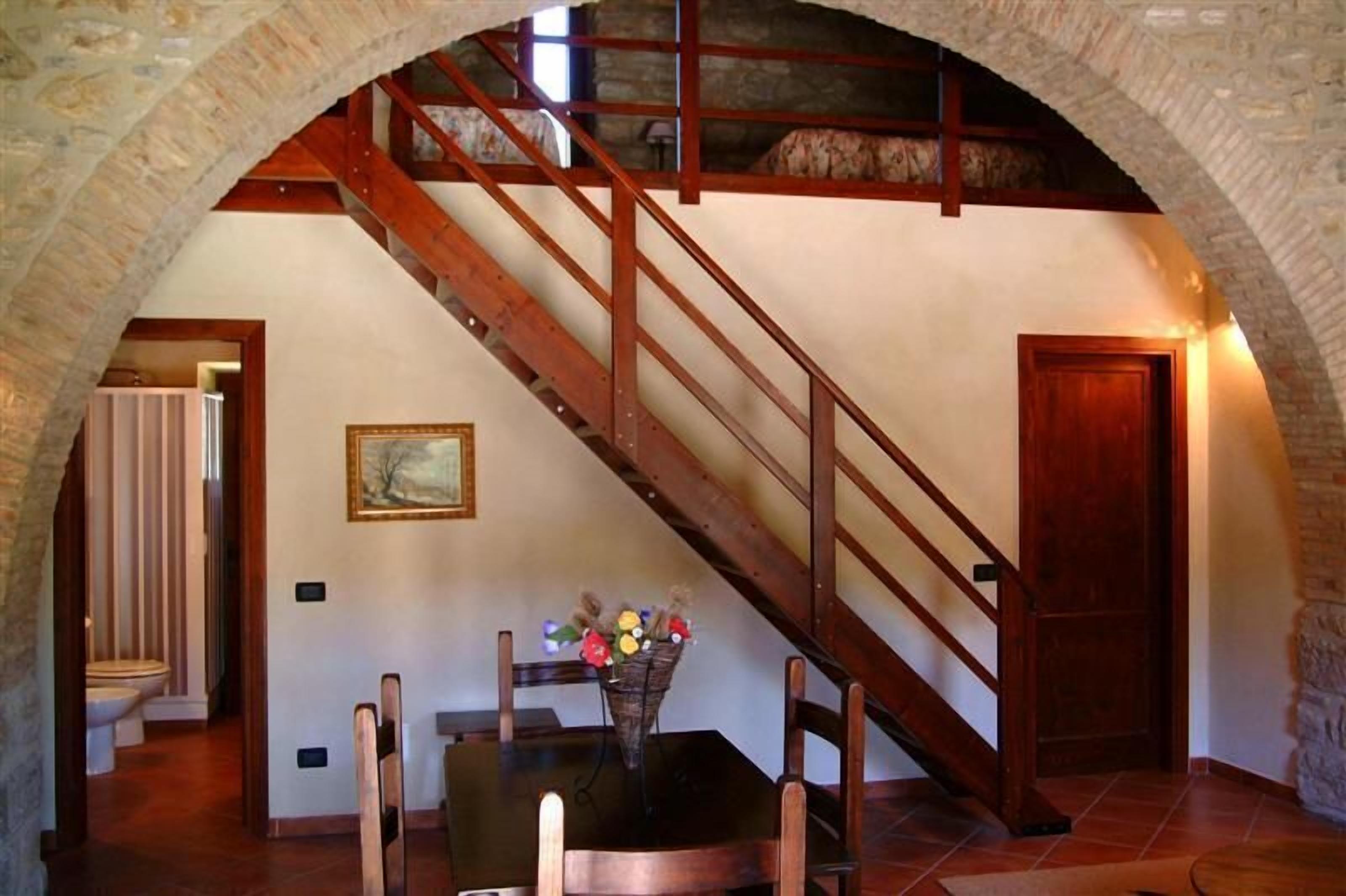Una escalera con barandillas de madera que sube a un nivel superior, una mesa de comedor con sillas y un jarrón de flores.