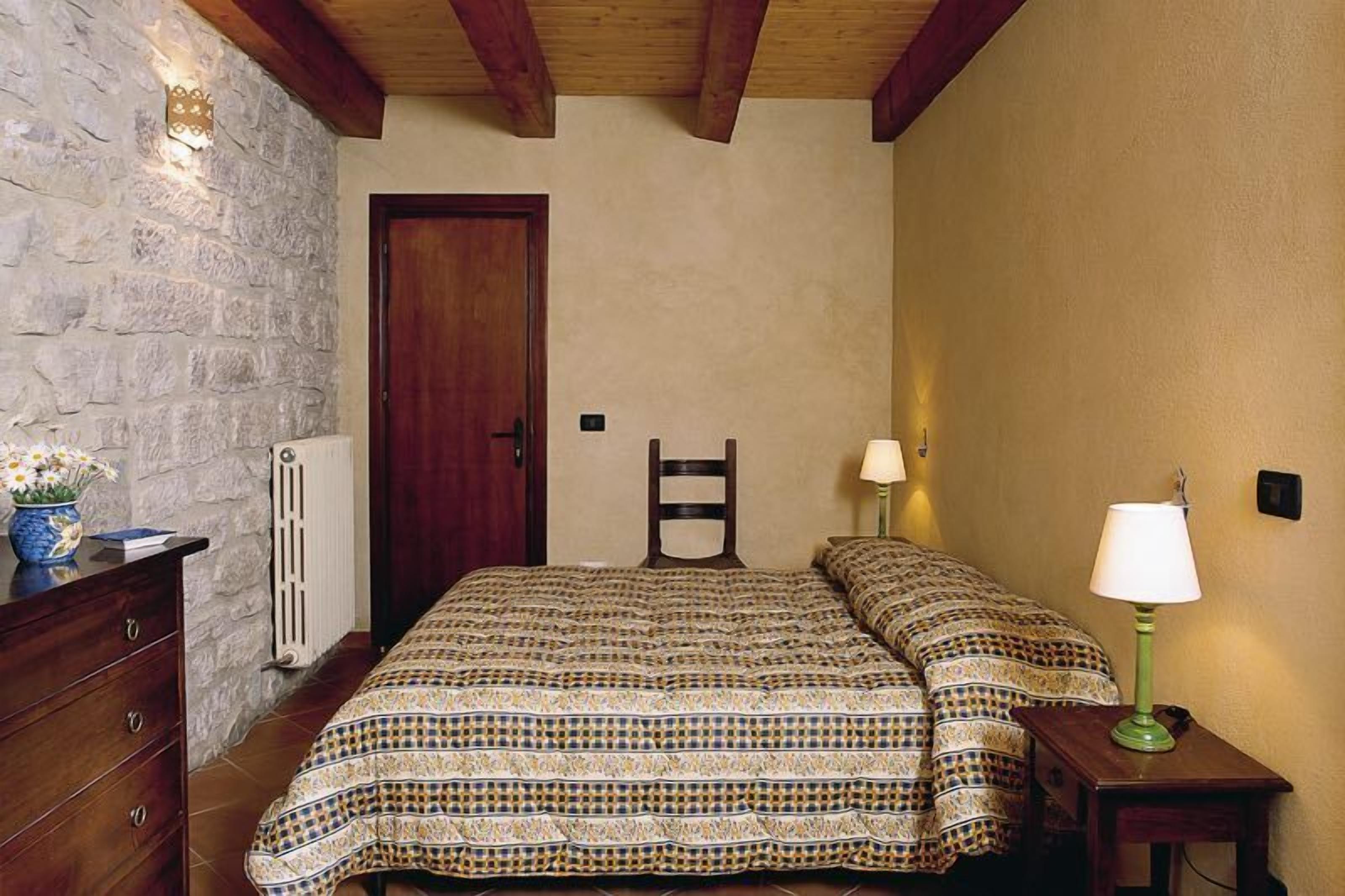 A bedroom with a patterned bedspread, a wooden dresser, a chair, and two bedside lamps.