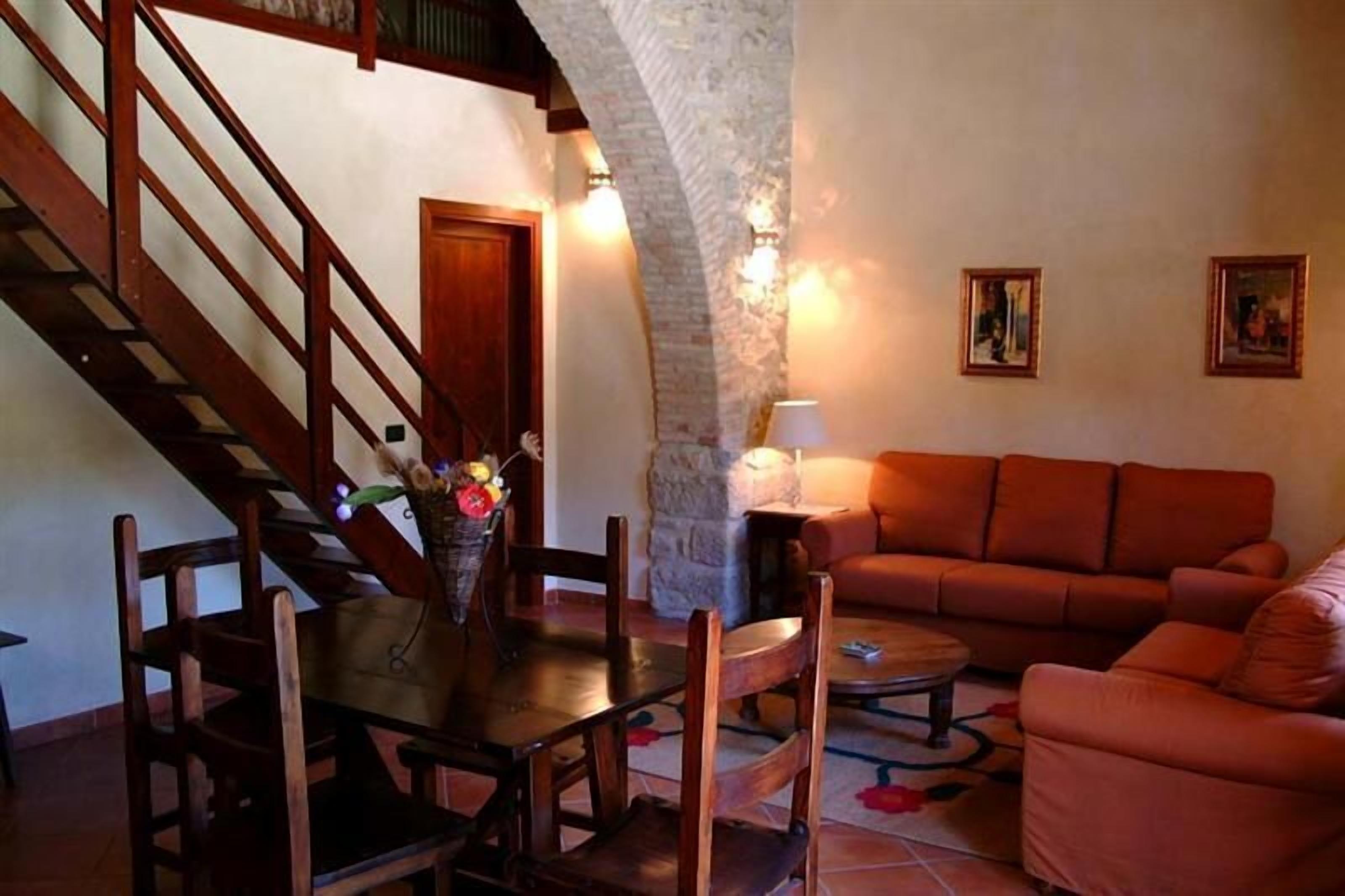 A cozy living room with a wooden staircase, a stone wall, a sofa, and a dining table with chairs.