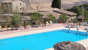 Un área de piscina con sillas de descanso y sombrillas, rodeada de edificios rústicos y un paisaje montañoso.