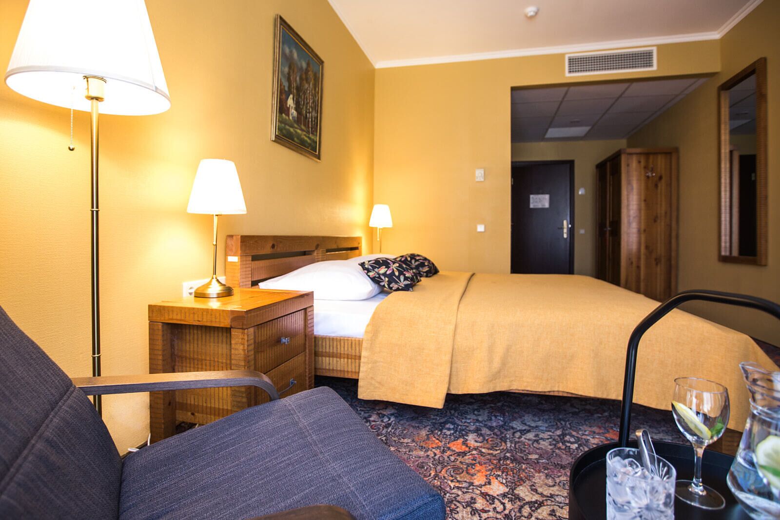 Habitación de hotel con cama, dos lámparas de noche, una mesita de noche, una silla, una pequeña mesa con vaso de agua y vaso de vino, y un cuadro en la pared.