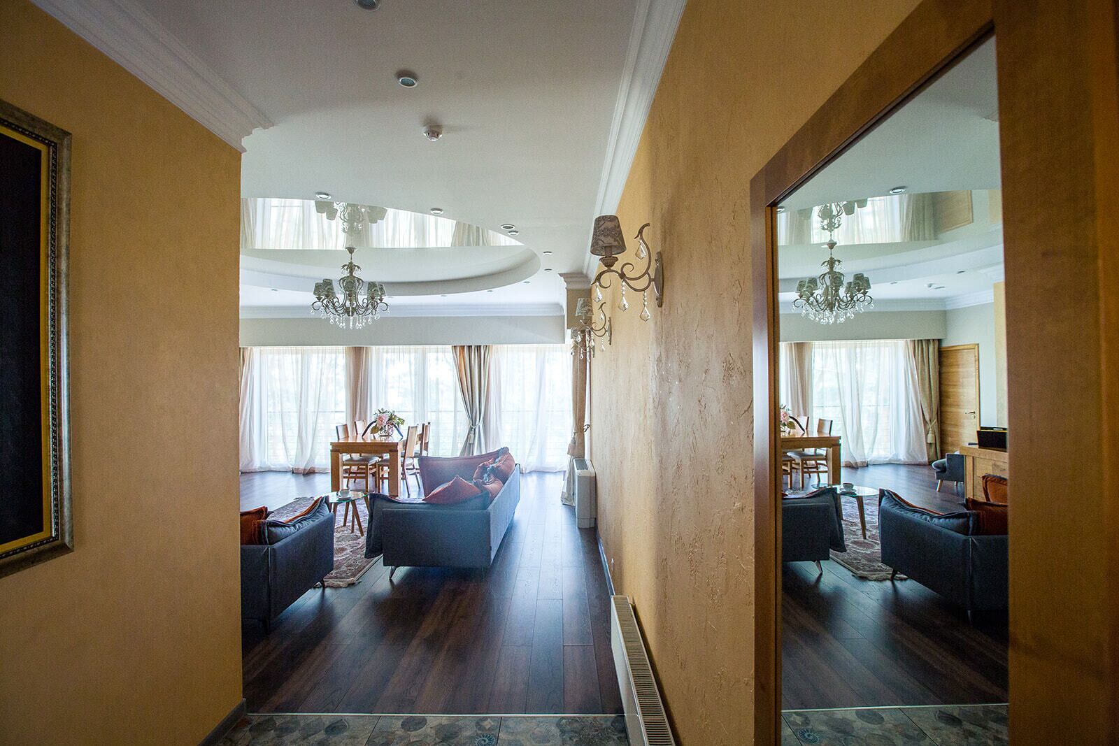 A spacious living room with a large mirror reflecting the room, a chandelier, and a dining area with a table and chairs.